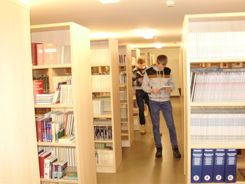 Skolas audzēkņi lasa bibliotēkā starp plauktiem
